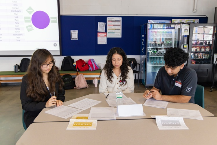 Three students seated at a table, reviewing papers, with a screen displaying a diagram behind them.