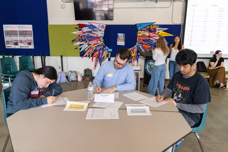 Four students at tables writing on papers. One woman sits at a table with a man.