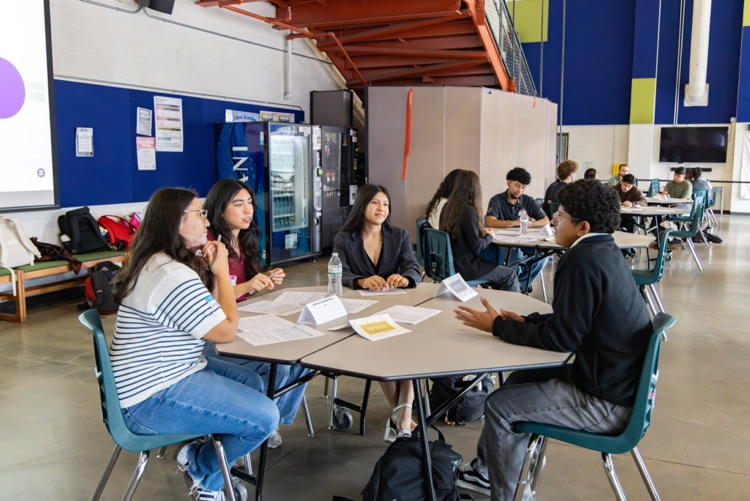 Students sit around a round table inside a classroom, discussing something with papers and a bottle.