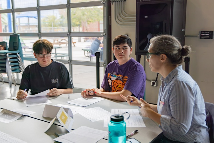 Three students seated around a table with papers and a water bottle, one holding a pen.