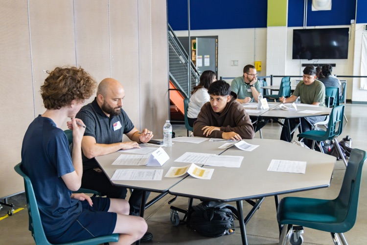 A group of people sit at tables inside a classroom. Papers and a water bottle on a table.