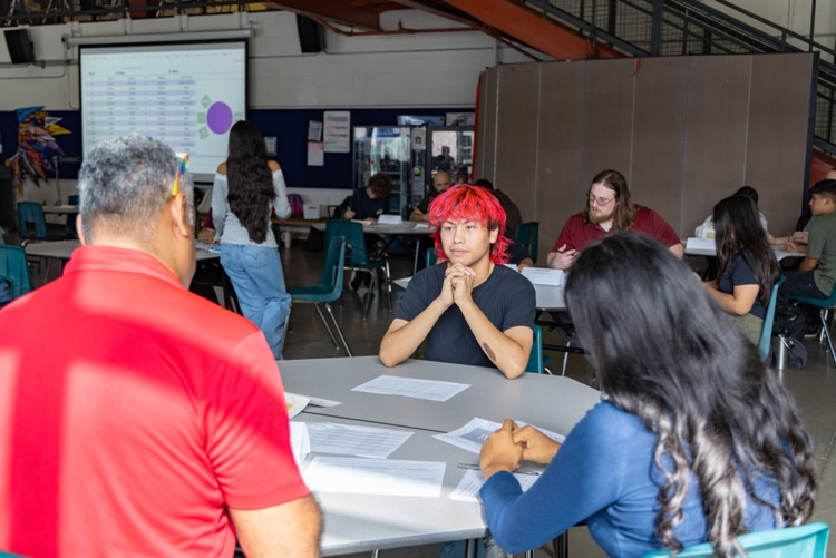 People sit at tables in a classroom. One person has red hair. Another person wears a red shirt.