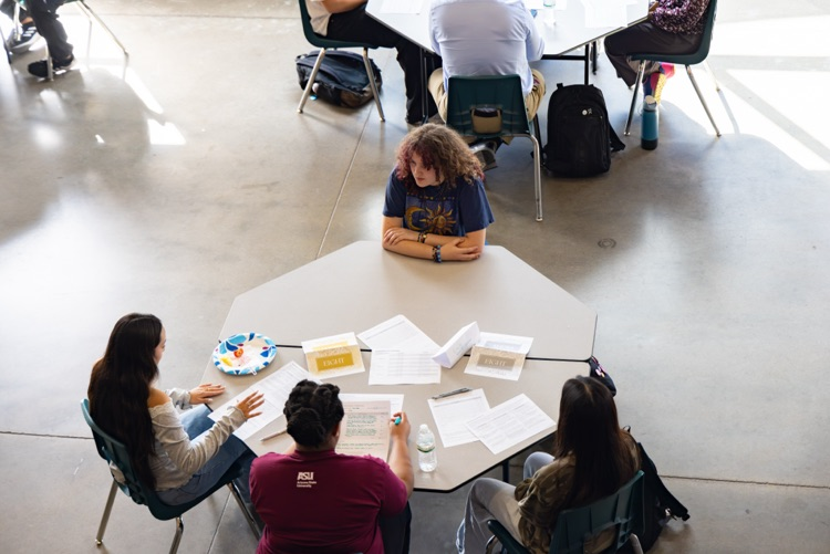 People sitting around a table with papers on it, some with backpacks nearby.