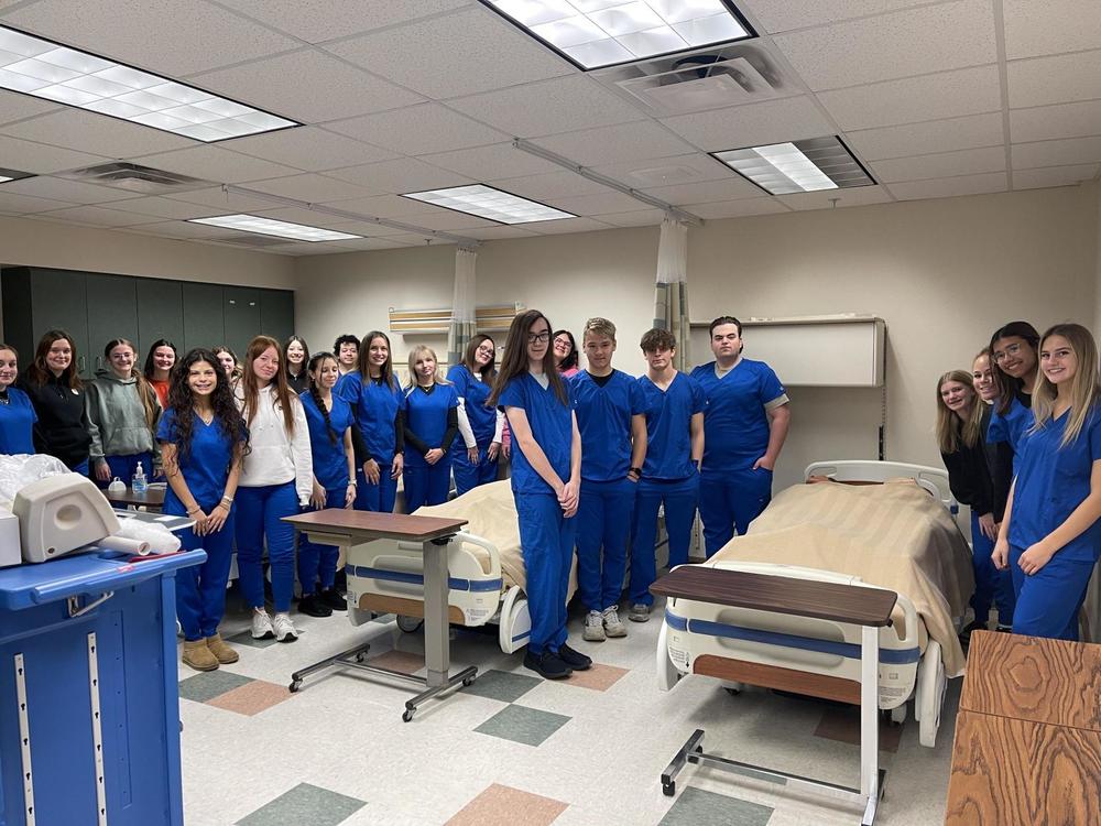 a group of students in blue medical scrubs stand surrounding hospital beds. 