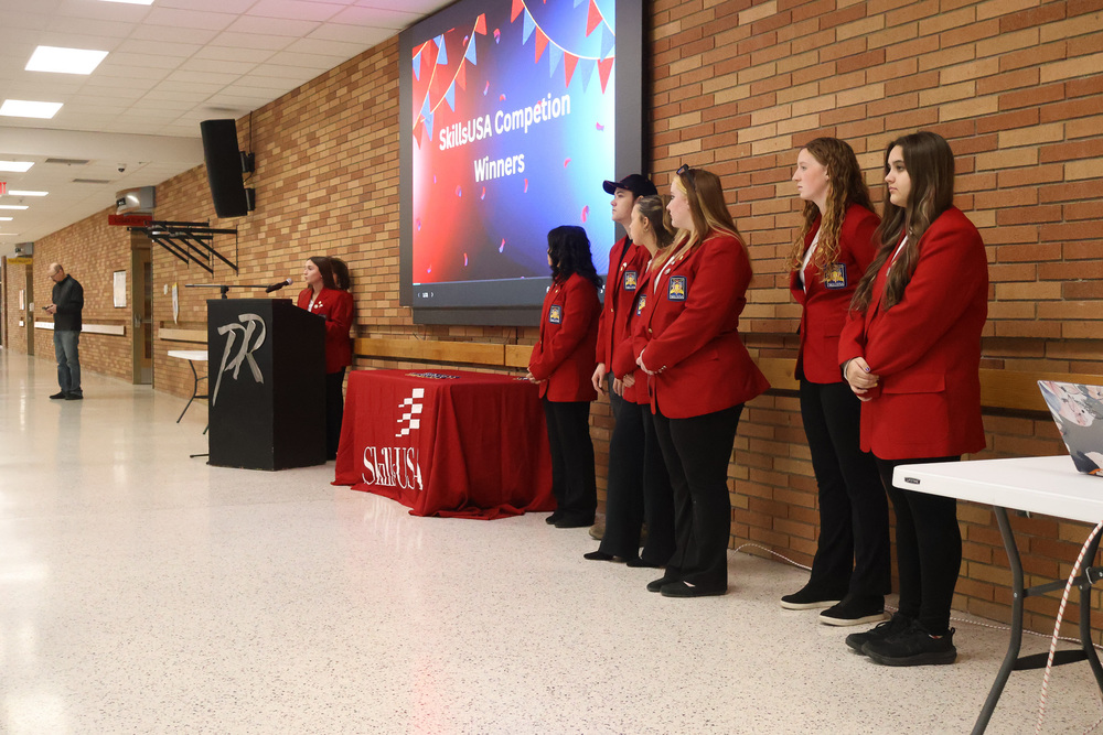 A group of male and female students stand in red jackets in front of a brick wall. There is one student at the podium speaking.