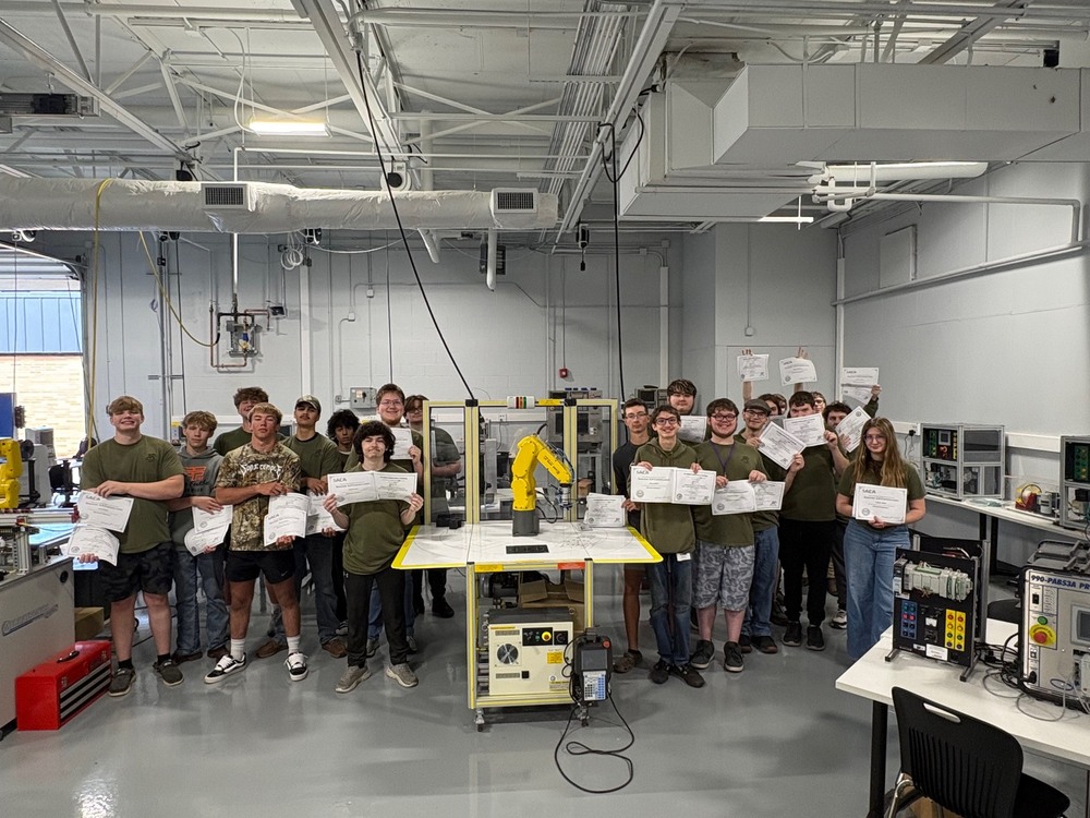 A group of students in dark green shirts stand around a yellow FANUC robot while holding certificates. 
