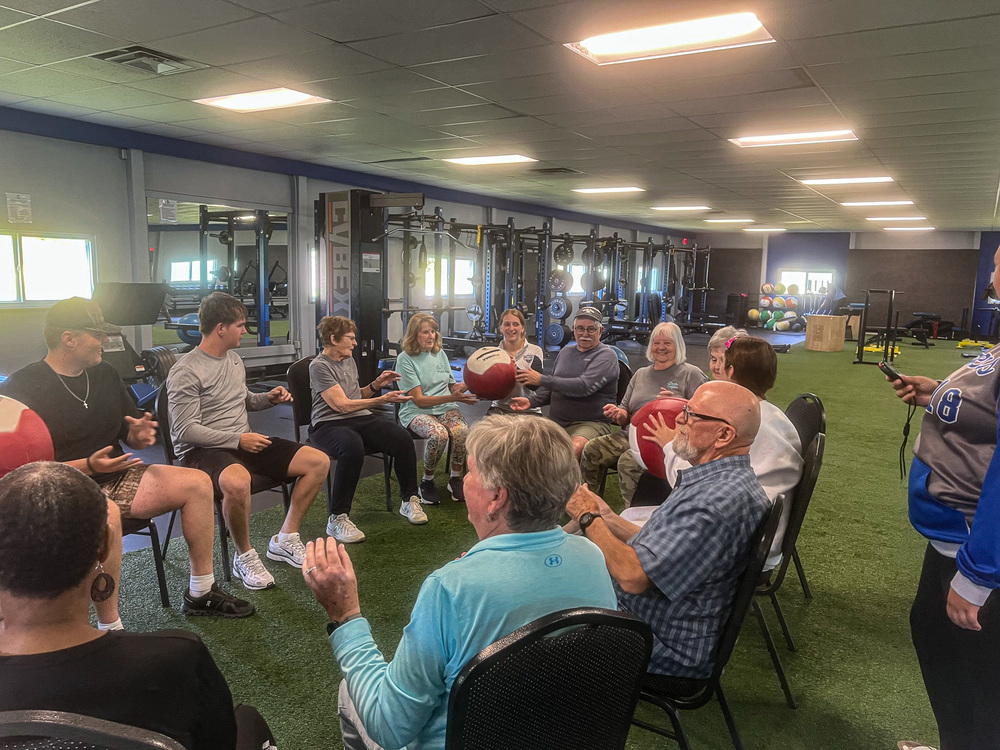 A group of students and senior citizens sit in a circle on black chairs while passing large balls around the circle.