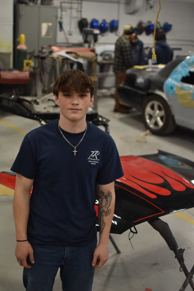 A teenage boy in a navy blue shirt with a PR logo on the front stands in front of a car hood on a stand. the hood is red with black flames. 