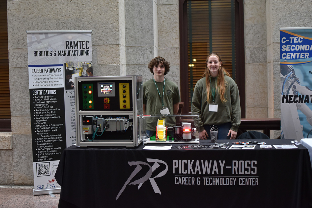 2 students stand behind a table with a small, cube like robot control system with a ferris wheel attached. One student is a female with red hair wearing a darker green sweatshirt and the other is a boy with medium length curly hair and a green shirt. The black table cloth reads Pickaway-Ross Career & Technology Center.