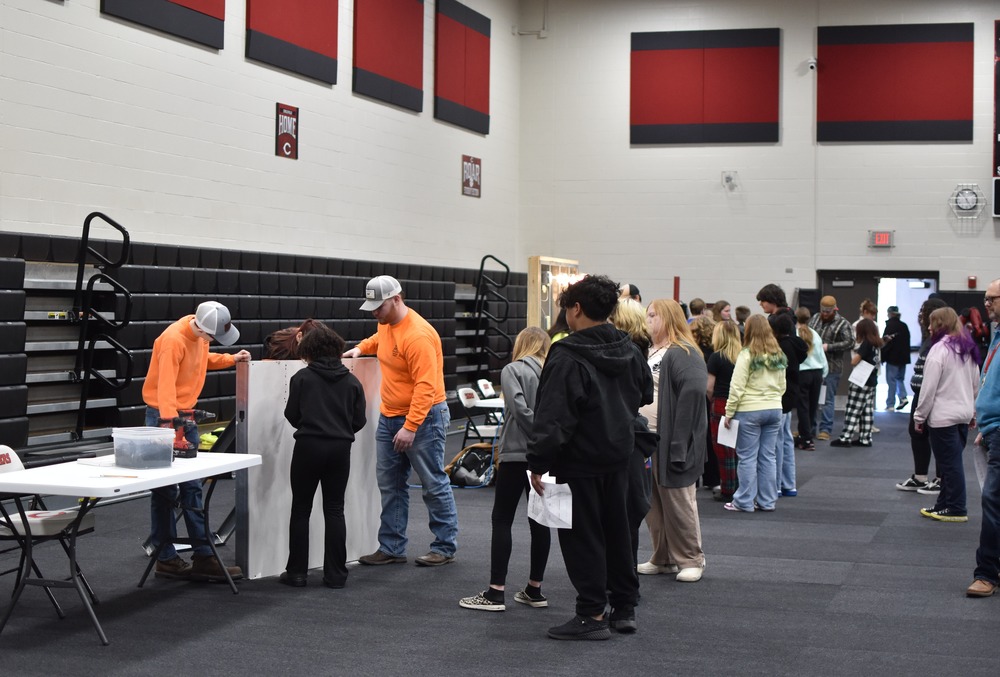 A group of 7th grade students, dressed in random apparel, watch students in orange shirts use a power tool. 