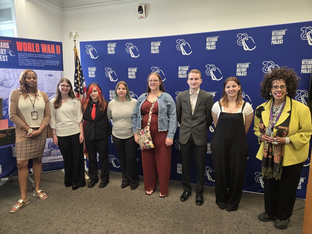 8 people stand in front of a banner that read Veterans History Project. 6 of them are our students who are dressed up. 
