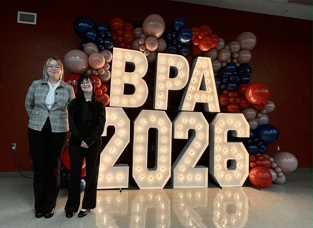 Two female students stand in front of a red white and blue balloon display. Inside the balloon arch, there are letters and numbers that read BPA 2026 and are lit up from within with multiple light bulbs. The students are dressed professionally, the student on the lefr has on black pants with a black and white plaid blazer and white under shirt. She has blonde hair pulled back with glasses. The student on the right is dressed in black pants and a black dress shirt. they have short black hair with bangs and glasses. 