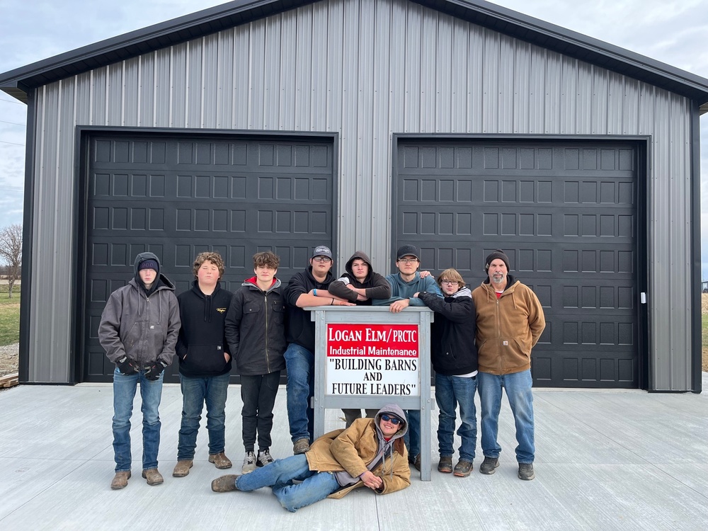 A group of students stands by a sign in front of a newly built pole barn that is gray and black. 