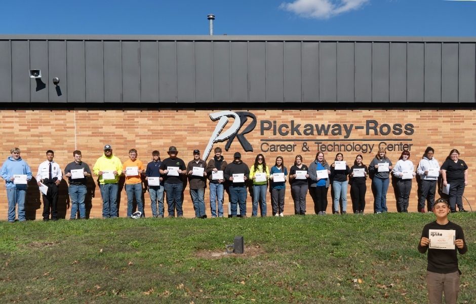 A group of students stand in front of a brick building a long row holding certificates. 