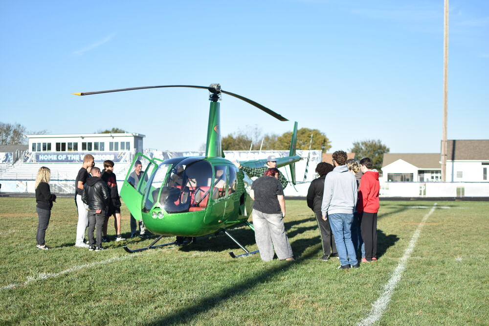 A group of students stand by a bright green helicopter on a green football field.