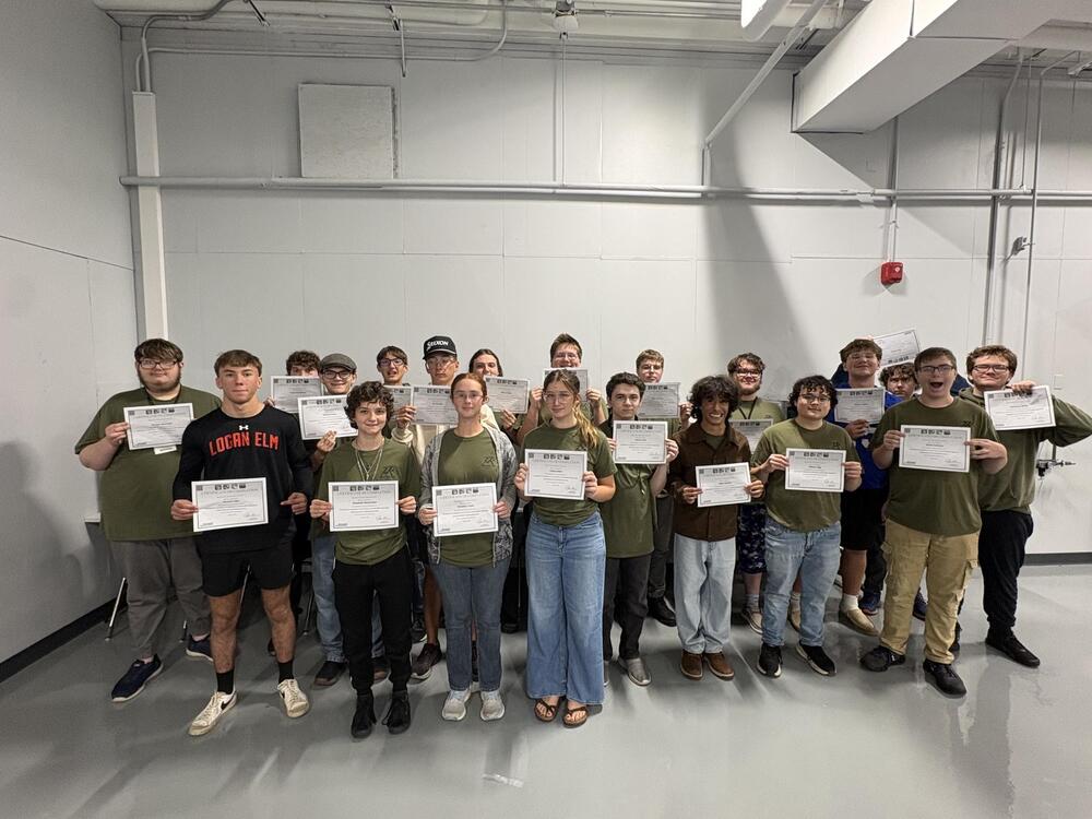 A group of students in dark green shirts hold certificates in front of them. 