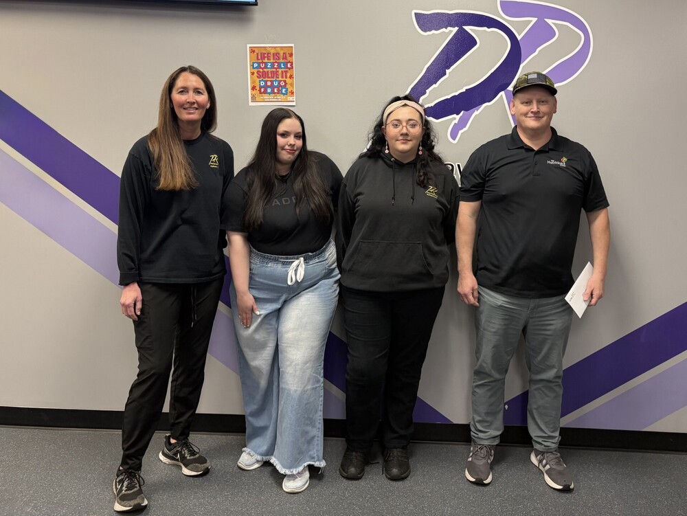4 people stand next to each other, all wearing black. A female teacher stands on the left side with 2 female students in between with a man in a black shirt and hat on the right.