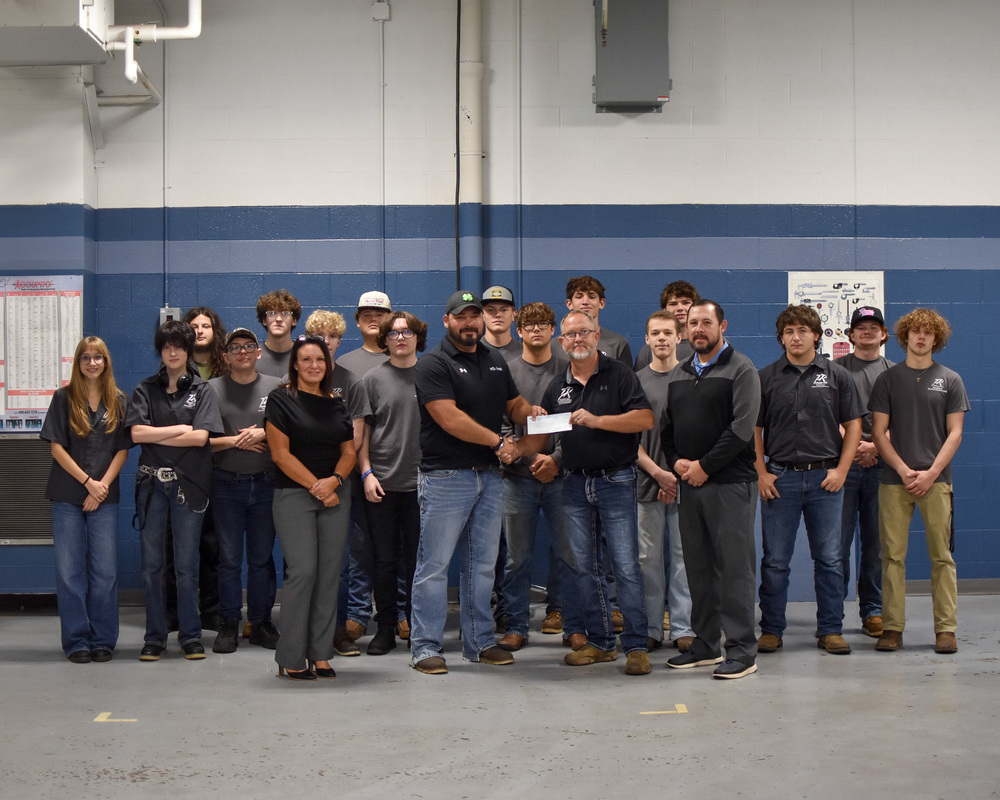 A group of students in gray work shirts stand in front of a white and blue wall in an industrial like setting. in front of them a man and woman in black shirts and jeans present a check to two men in black shirts.