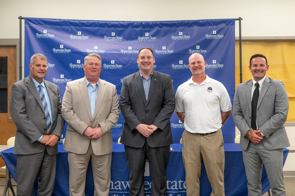 A group of men dressed in suits stand in front of a blue background with the word logo of Shawnee State University all over it.