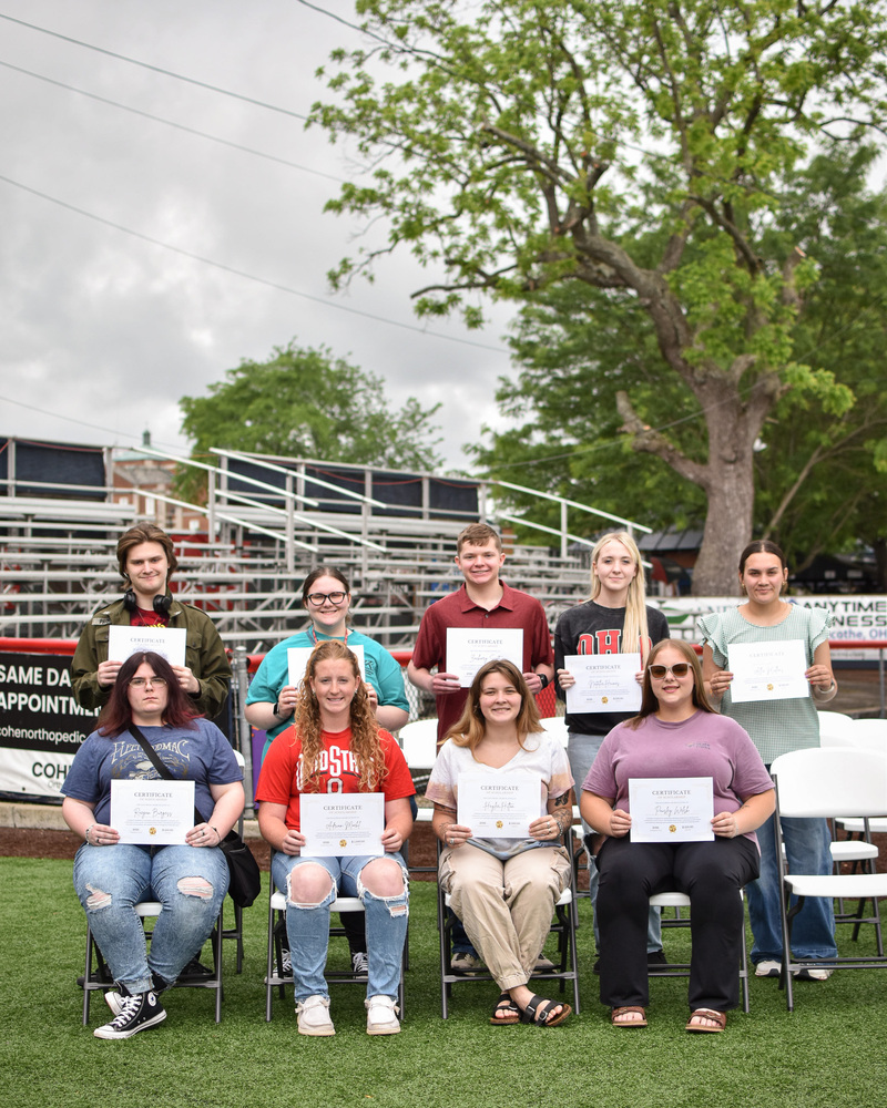 A group of students sit in chairs with students standing behind. 4 are sitting while 5 are standing. They are all holding white certificates. 