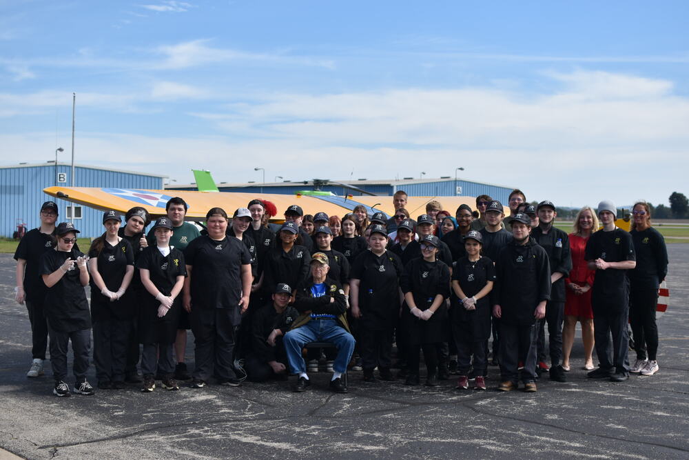 A group of students stand behind an older man seated in a chair. they are all standing in front of a plane.