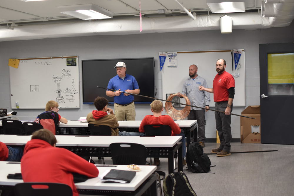3 men stand in front of a classroom. One is holding a long metal pipe and one is holding copper wire