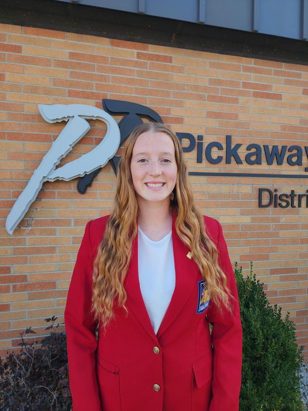 A girl with red hair stands in front of a brick wall with the Pickaway-Ross logo in the background. She is wearing a red jacket with a white undershirt. She is smiling. 