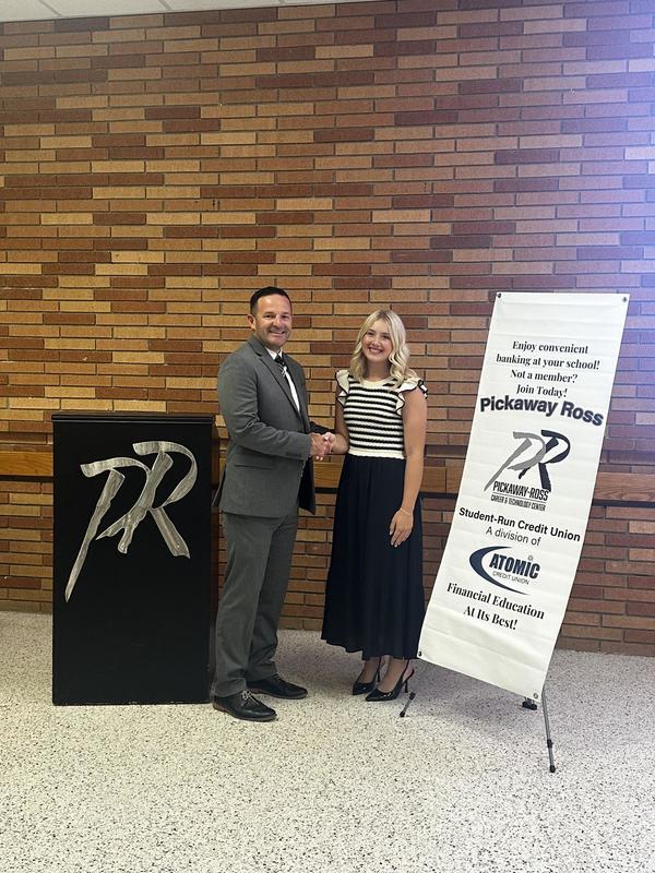 A man in a gray suit shakes the hand of a female student in a white and black striped top with a black skirt. They stand in front of a brick wall and are flanked by a black podium with a PR on it and a white sign with tiny words. 