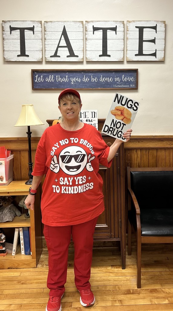 School nurse standing in front of a school sign holding a sign that reads NUGS NOT DRUGS. She is wearing red for red ribbon week,
