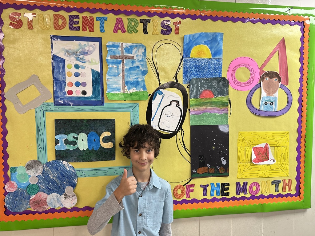 Male student standing in front of student decorated  bulletin board holding a thumbs up