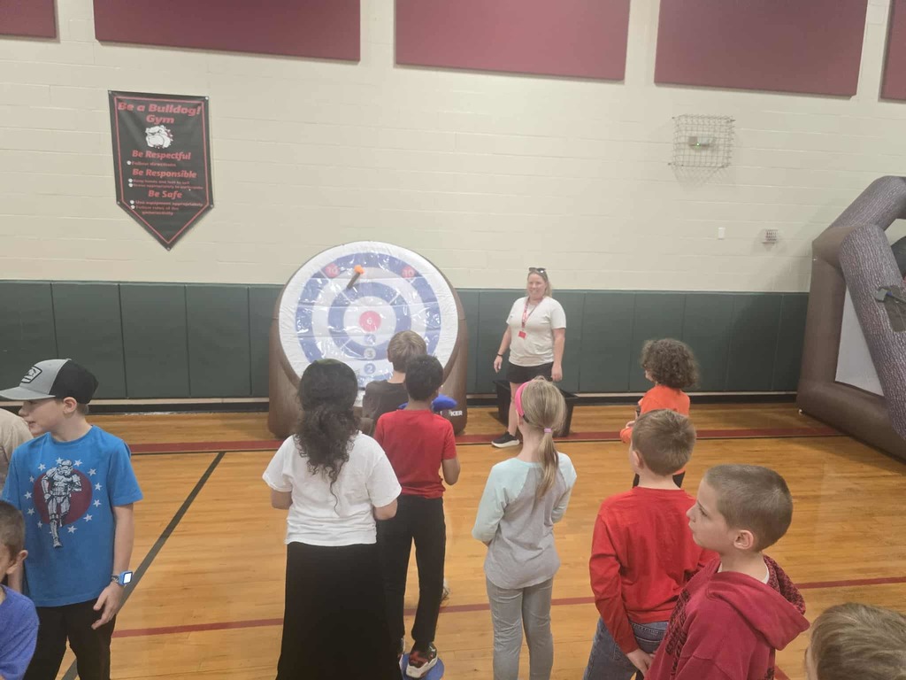 Group of kids standing in a row in a school gym throwing darts at an inflatable dartboard