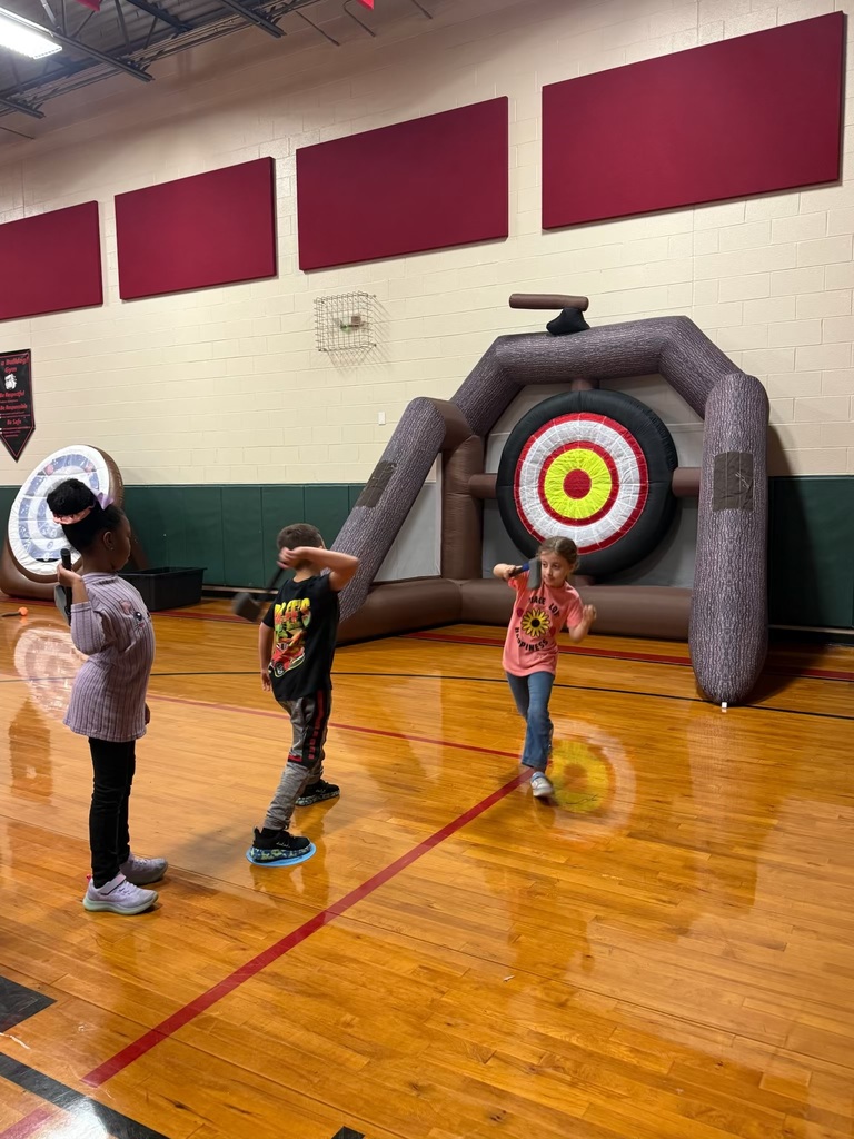 Three kids standing in a row in a school gym throwing a toy ax at an inflatable bullseye
