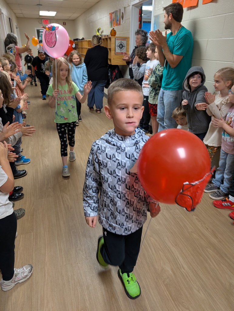 Group of kids walking in a school hallway holding balloons on sticks with kids lined up standing on both sides of the hallway