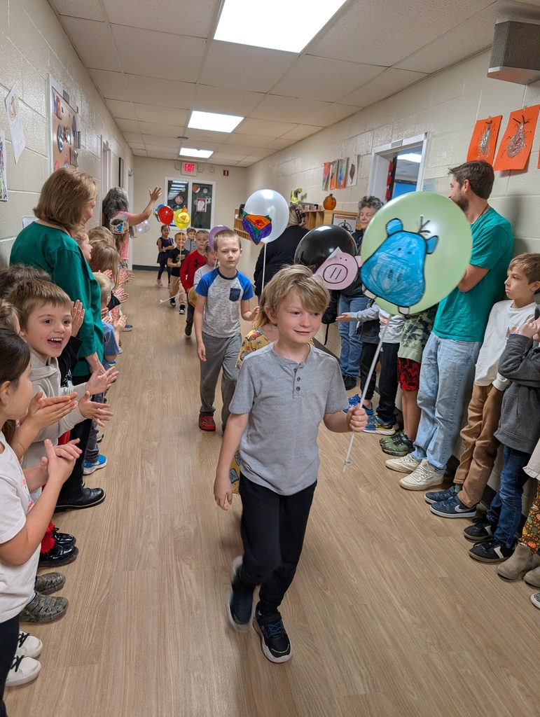 Group of kids walking in a school hallway holding balloons on sticks with kids lined up standing on both sides of the hallway