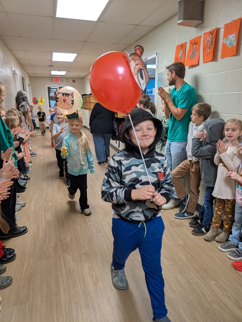 Group of kids walking in a school hallway holding balloons on sticks with kids lined up standing on both sides of the hallway