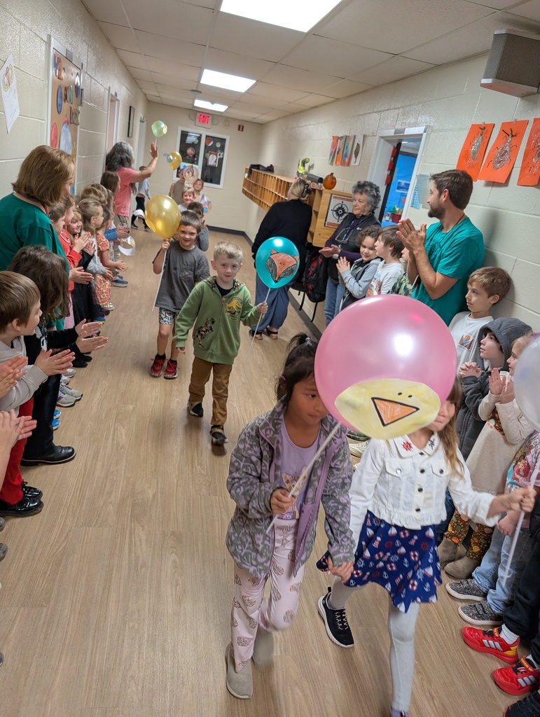 Group of kids walking in a school hallway holding balloons on sticks with kids lined up standing on both sides of the hallway
