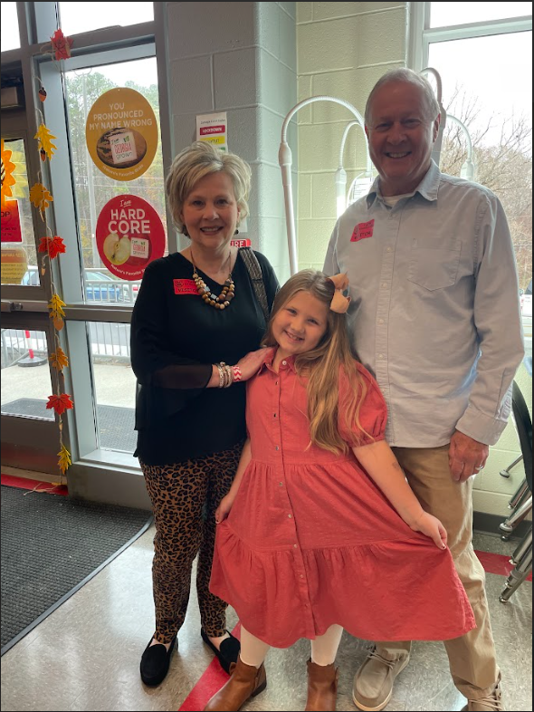 two grandparents standing behind a female child in the school cafeteria standing and smiling 