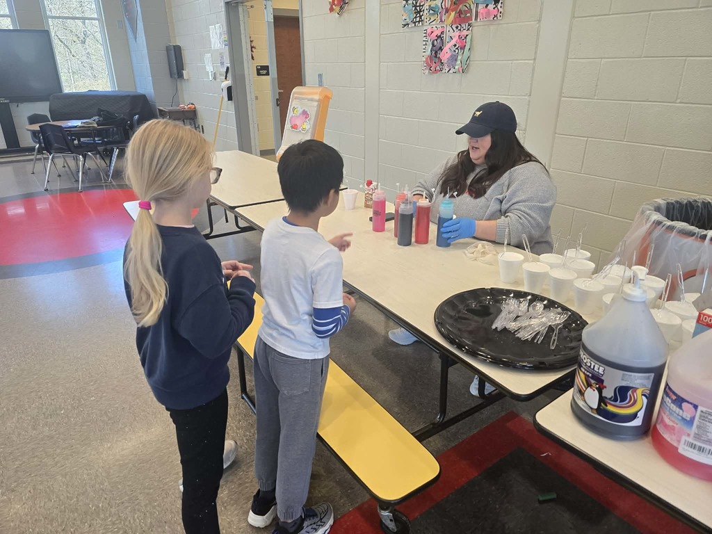 Two kids in line in front of a school cafeteria table. Lady sitting behind table with bottles of snow cone flavors