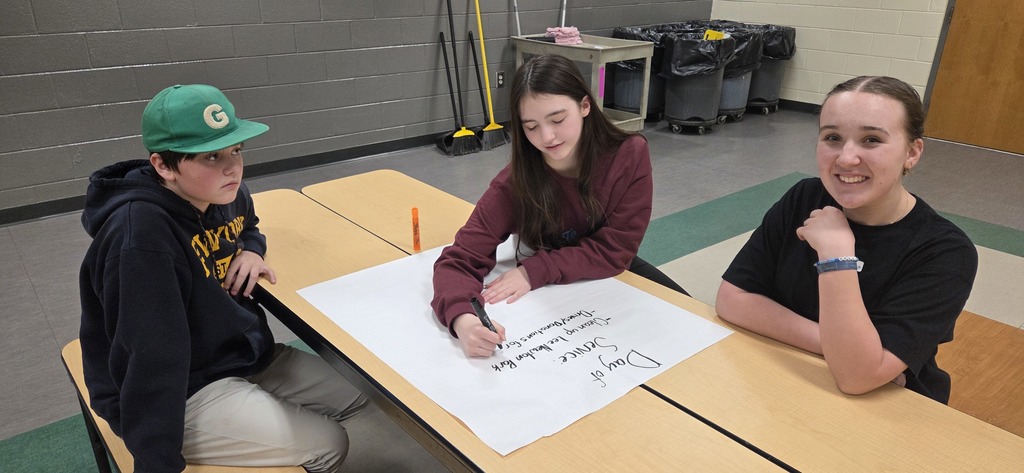 3 students sitting at cafeteria table. One is writing on chart paper.