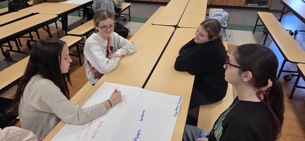 4 students sitting at table. One is writing on chart paper.