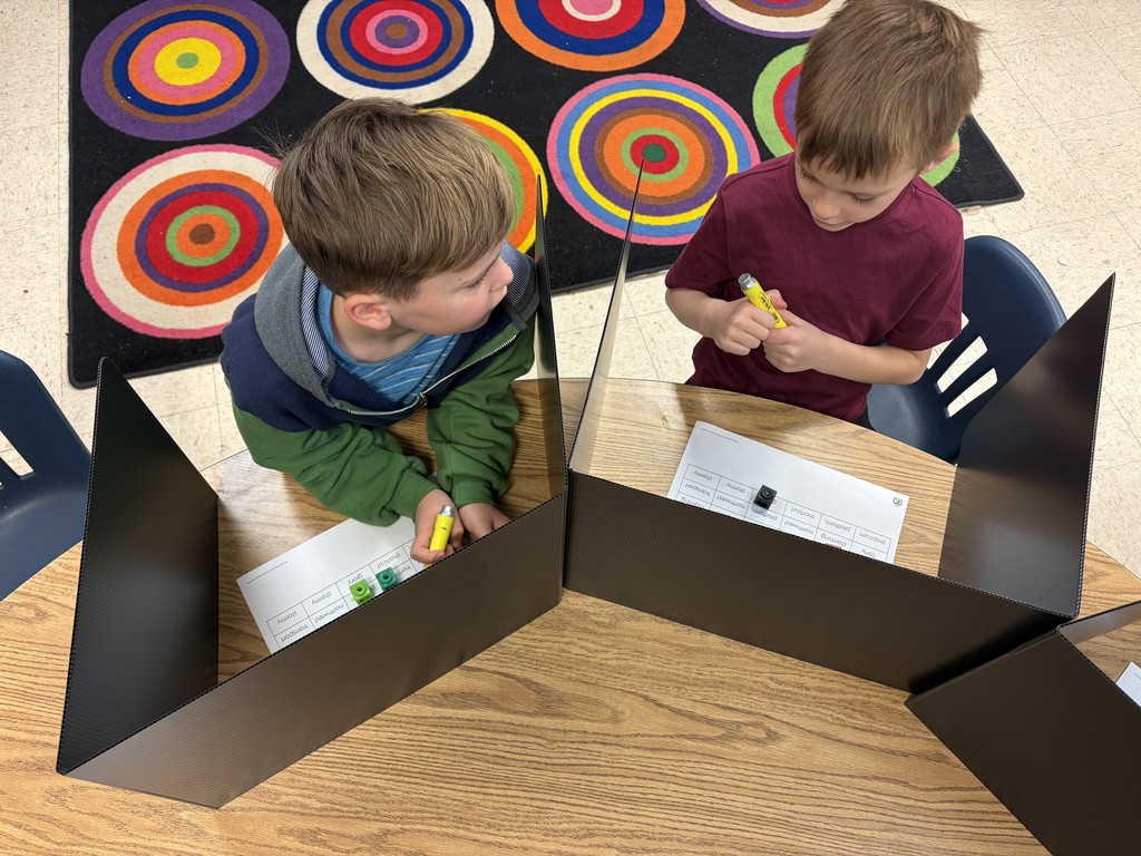 Two students  sitting at a table with black dividers playing a dice game both holding yellow highlighters