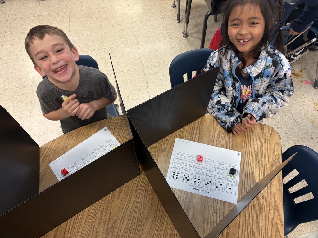 Two students  sitting at a table with black dividers playing a dice game