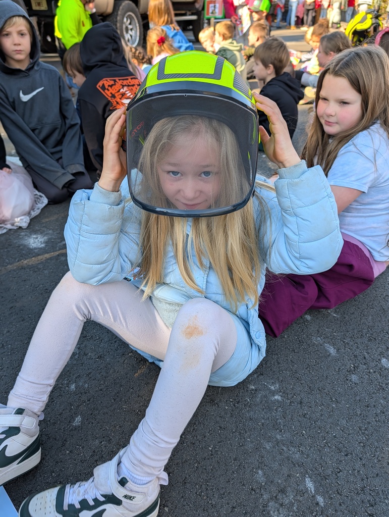 classroom of students sitting on the ground, one female student is wearing a construction hat