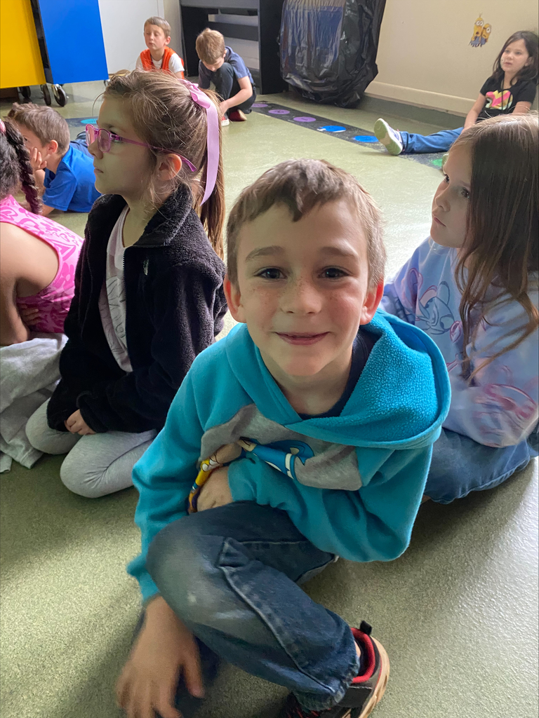 Boy sitting on the floor. He is smiling at the camera  and there are other children sitting around him.