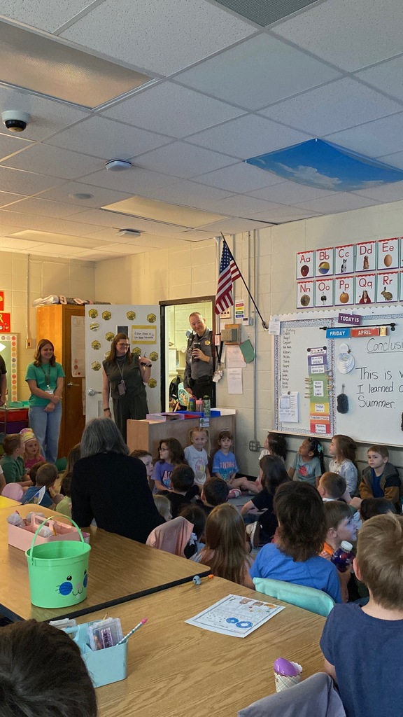 classroom of students sitting on the ground, adults standing in a classroom