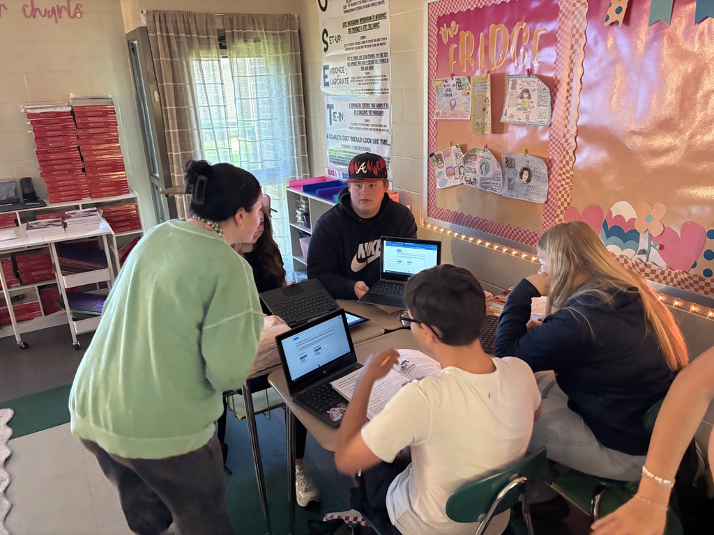 Group of students seated at desks working. Mrs Sutton is checking on them.