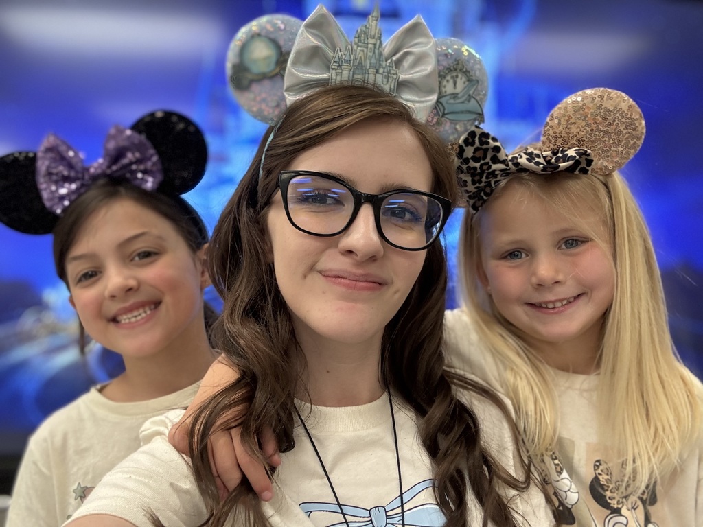 Two girls and a woman wearing minnie mouse ears. They are standing in front of Cinderella's castle projected on a screen.