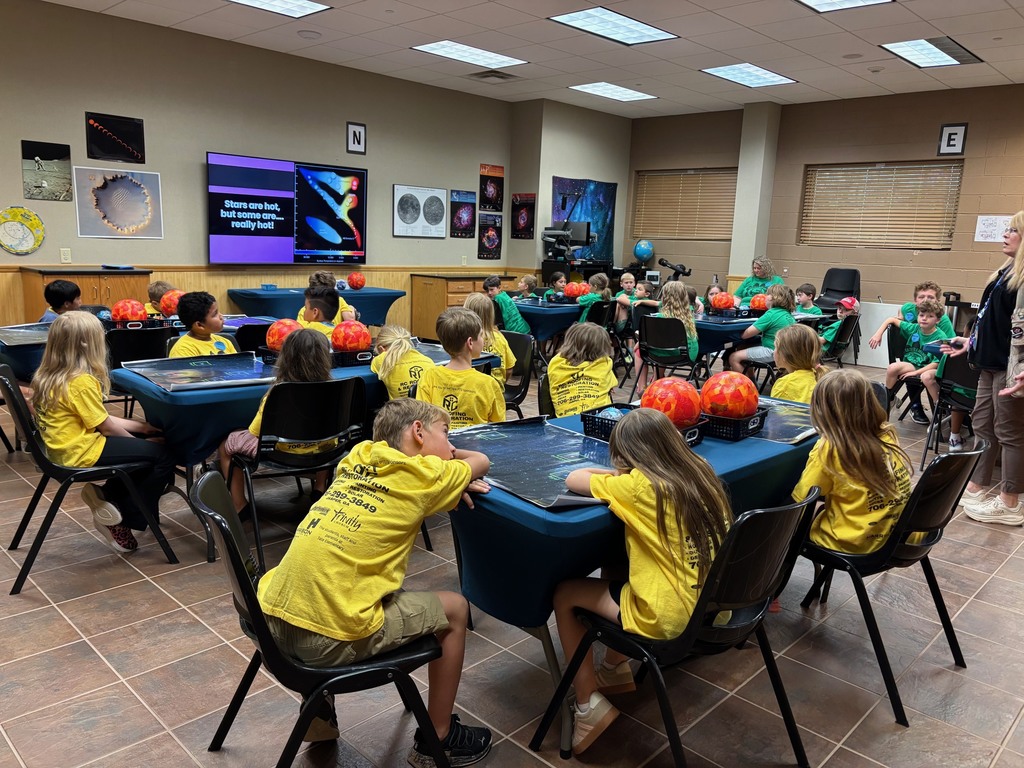 Large group of kids sitting at tables some are wearing green shirts, some are wearing yellow shirts