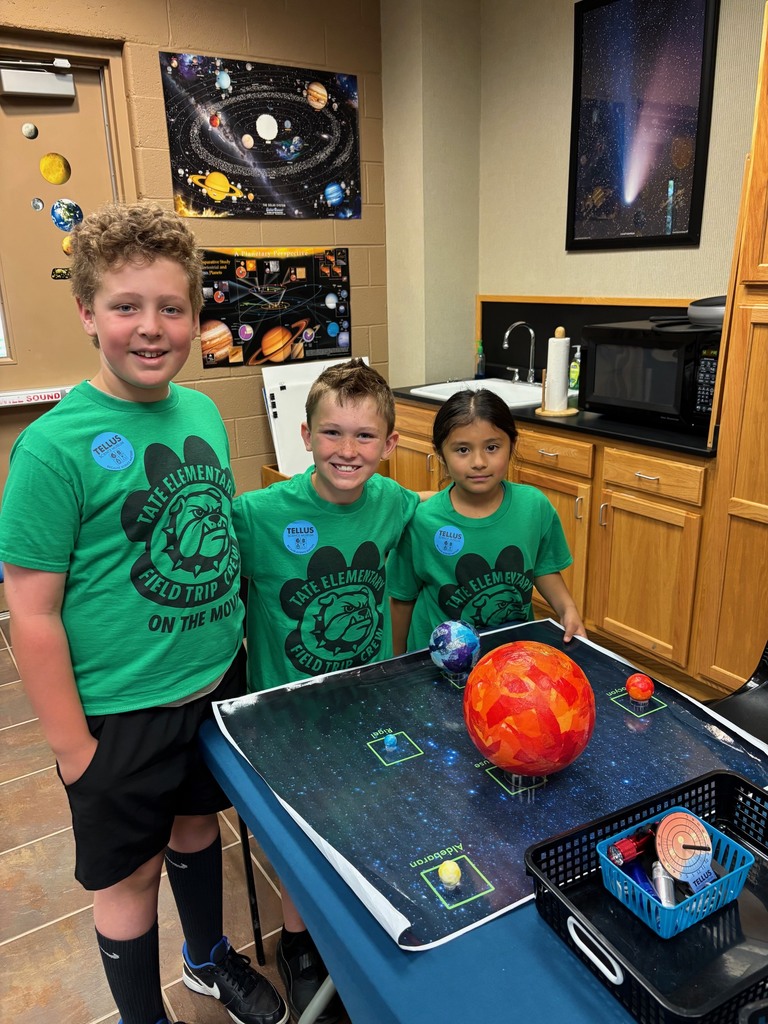 Three students in green shirts standing around a group of planets