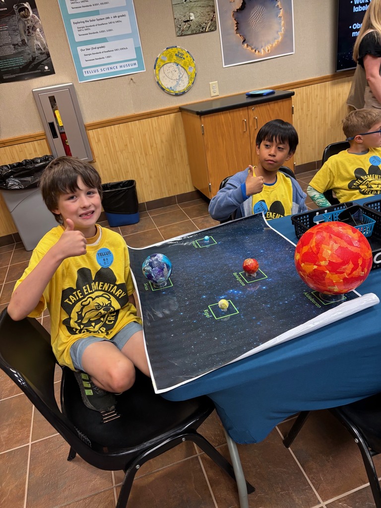 Three students in yellow shirts sitting at a table around a group of planets